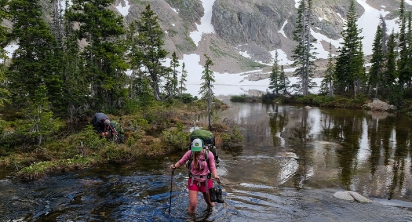 A person wearing a backpack uses a trekking pole as they walk through a shallow alpine lake.